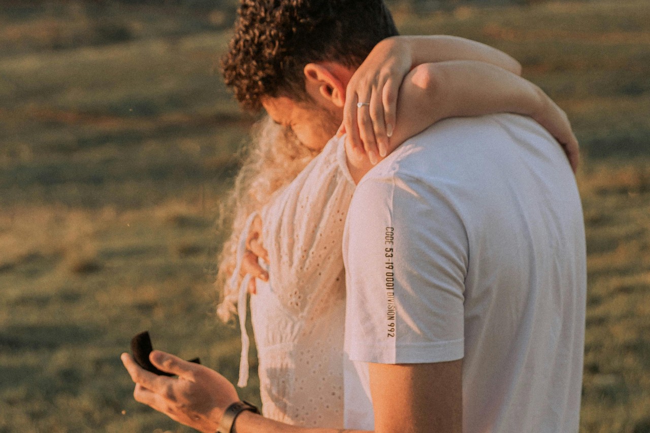 A couple shares a heartfelt embrace in a sunlit field as an engagement ring box is held, capturing a romantic proposal moment.