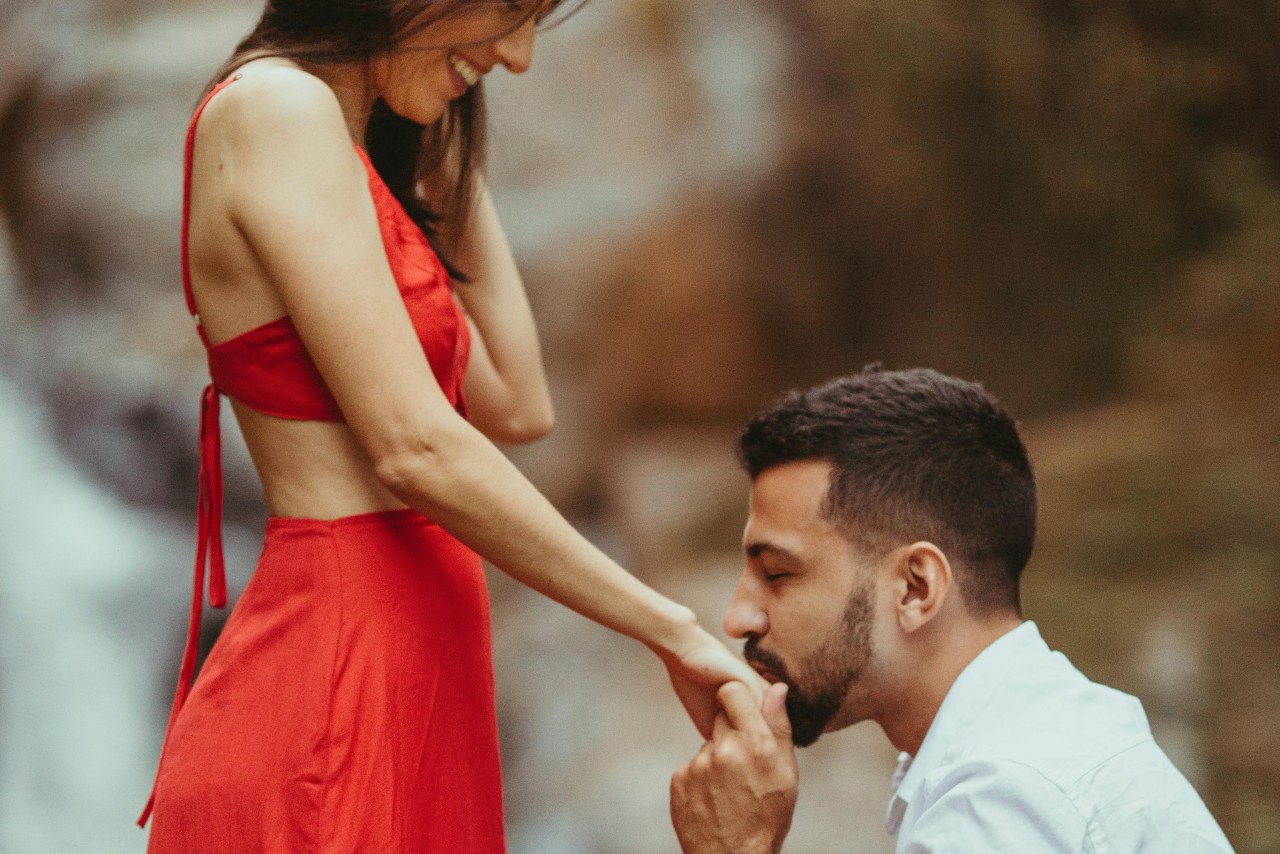 A man kneels, proposing to a woman in red dress against a blurred background.