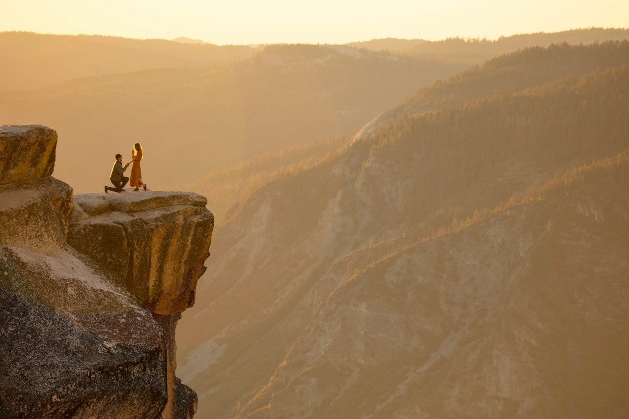 A person kneels for a marriage proposal on a cliff edge at sunset.