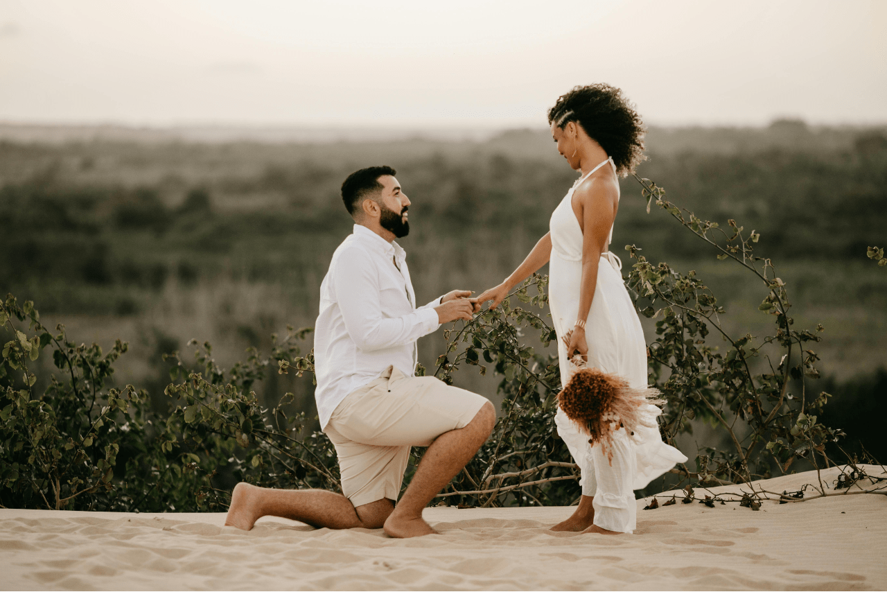 A man kneels and proposes to a woman holding flowers on a sandy hill, with greenery and a blurred landscape in the background.