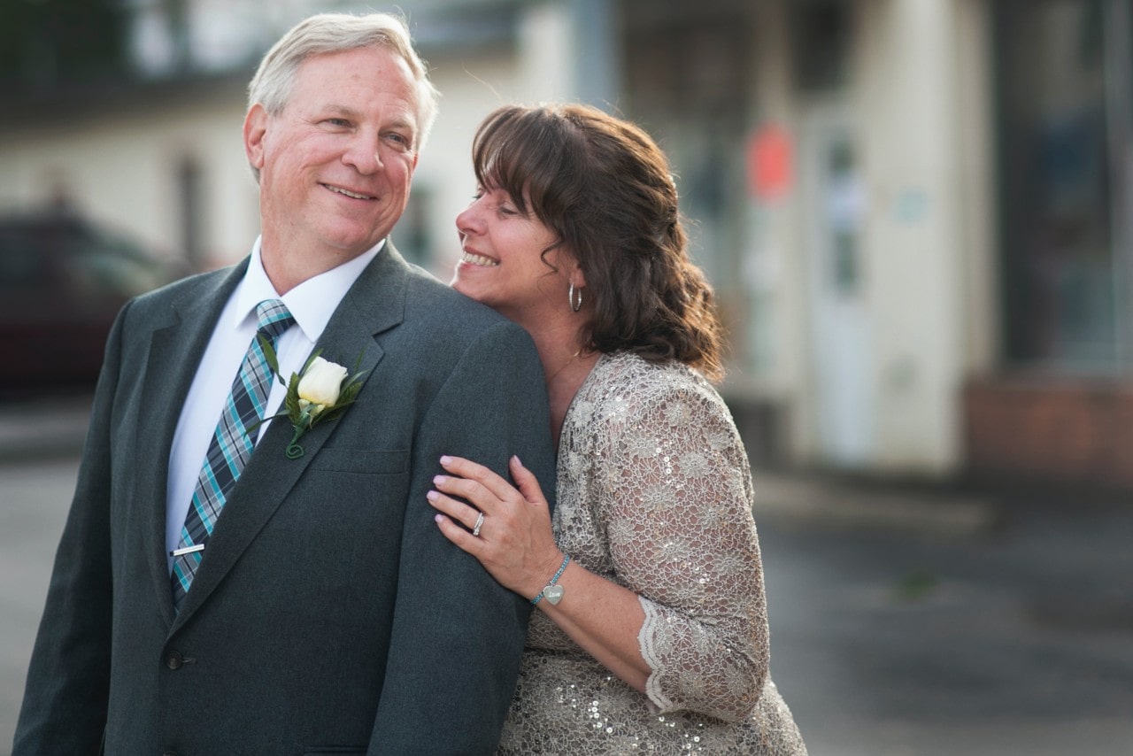 An older couple smiles warmly; the man in a suit with a floral boutonniere, the woman in a sparkling dress wearing elegant jewelry, leans affectionately on his shoulder.