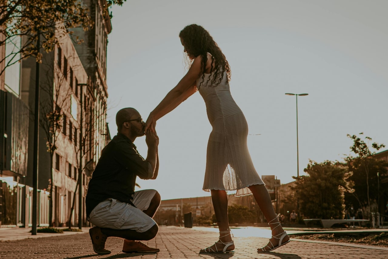 A man down on his knee, kissing his bride-to-be&rsquo;s hand as they stand on a city strip sidewalk.