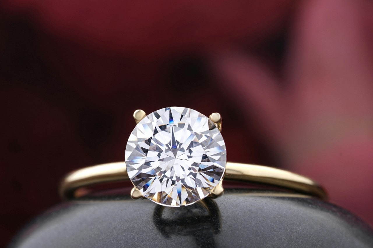A close-up image of a yellow gold solitaire engagement ring with a rounded center stone, sitting on a black reflective ball with a deep red blurred background.