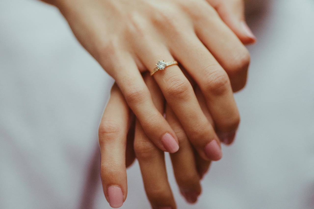 A woman with painted pink nails, resting her hands on top of one another, showcasing her yellow gold solitaire engagement ring.