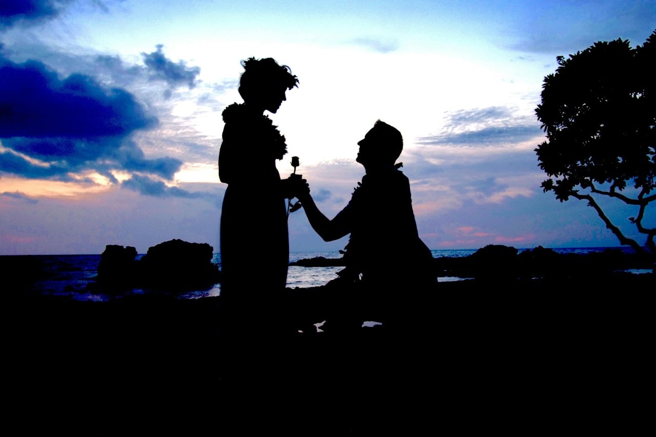 A back lit photo with the shadow of a man proposing to his bride-to-be, in front of a beautiful view of a body of water at sunset.