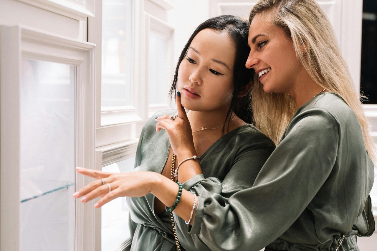 Two women twinning in olive green outfits examine a jewelry display case.