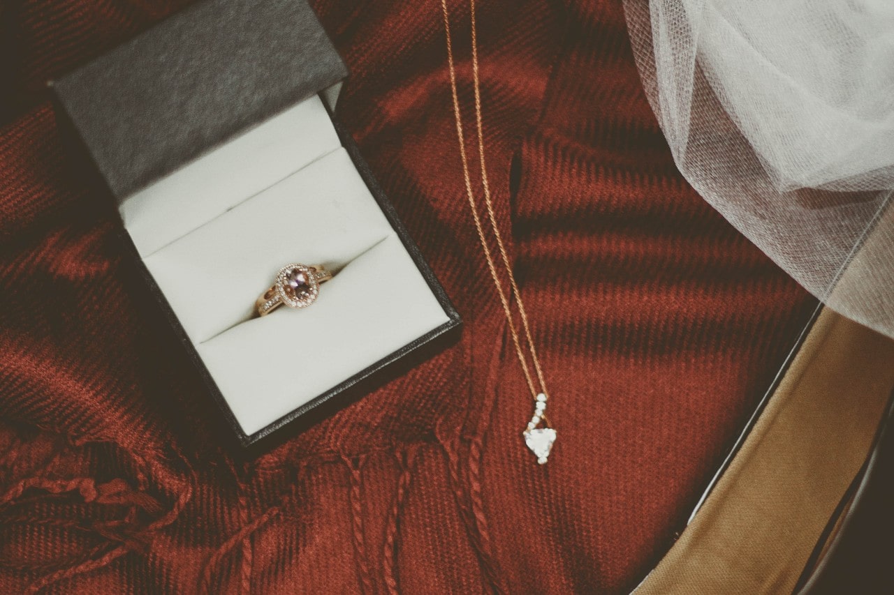 A delicate ring with a pink stone in a black box is set on a rich red fabric. Beside it is a gold necklace with a sparkling pendant.