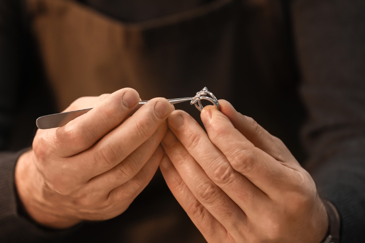 A close up of a jeweler&rsquo;s hands inspecting a diamond engagement ring using a metal tool.