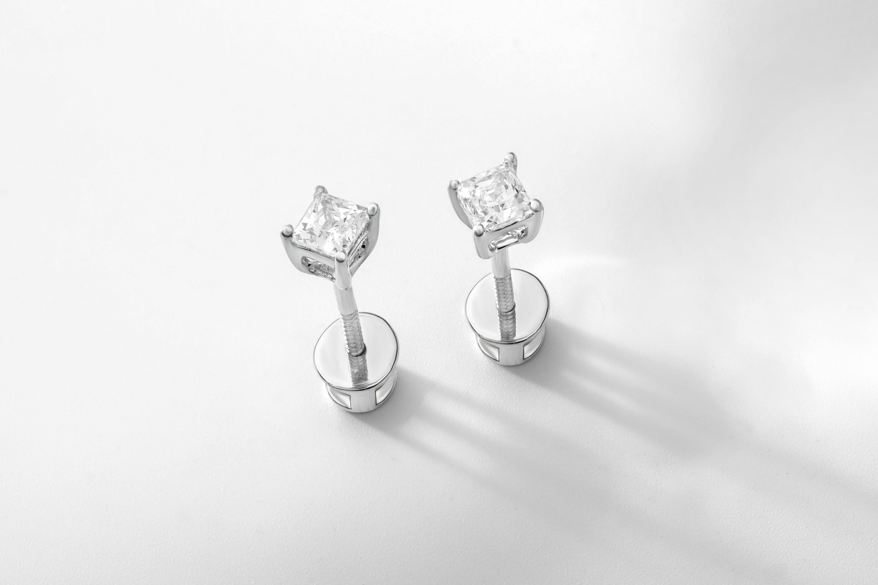 A close-up of a pair of princess cut diamond stud earrings on a white surface.