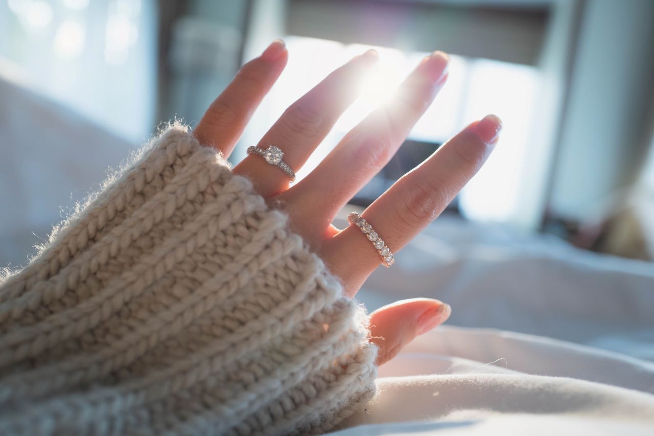 A close up of a hand in a white knitted sweater against the sunlight, adorned with white gold diamond engagement ring and wedding band.