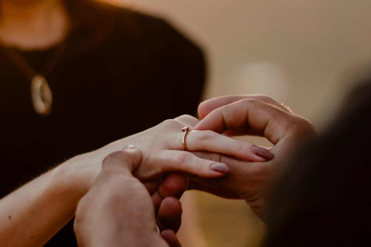A man putting a golden engagement ring on a woman’s finger with a blurred romantic background.