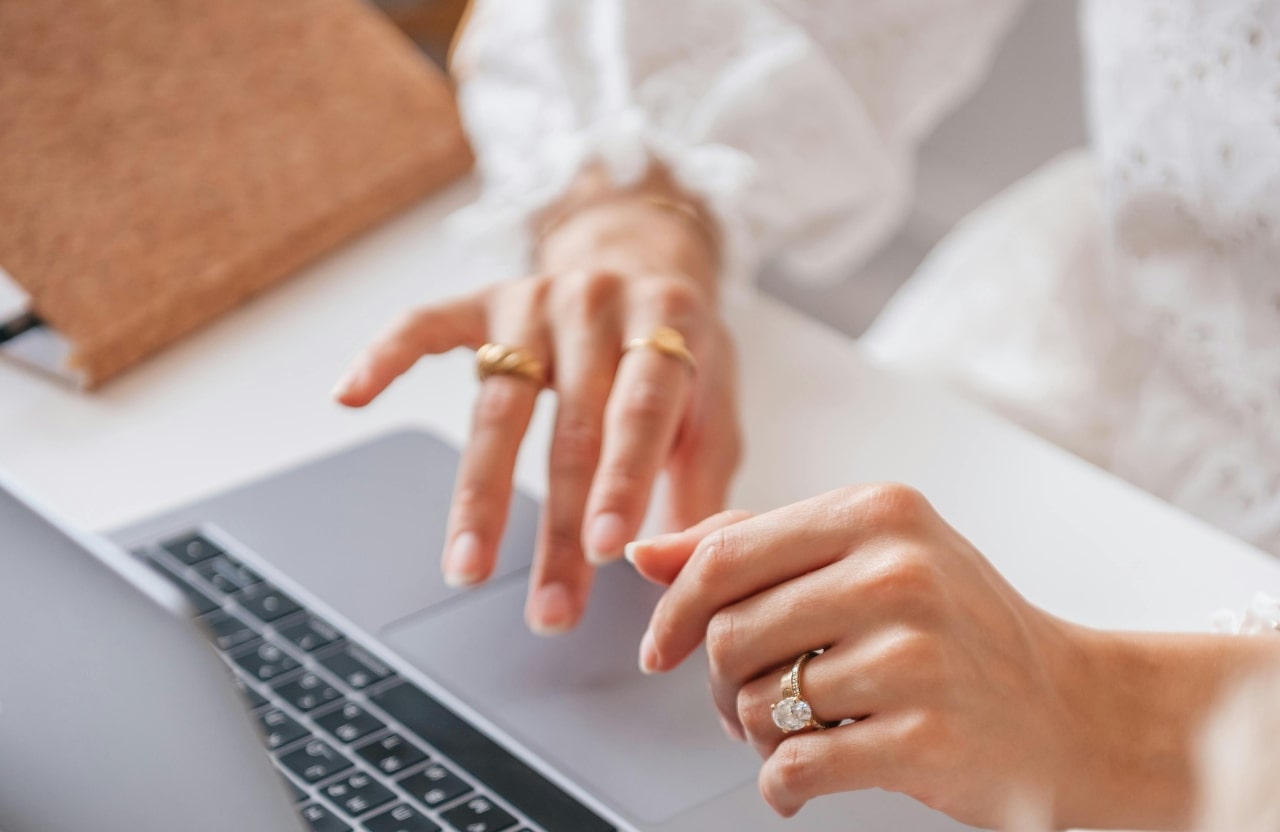 An elevated view of a woman’s hand typing on a laptop, with focus on her oval cut solitaire engagement ring.
