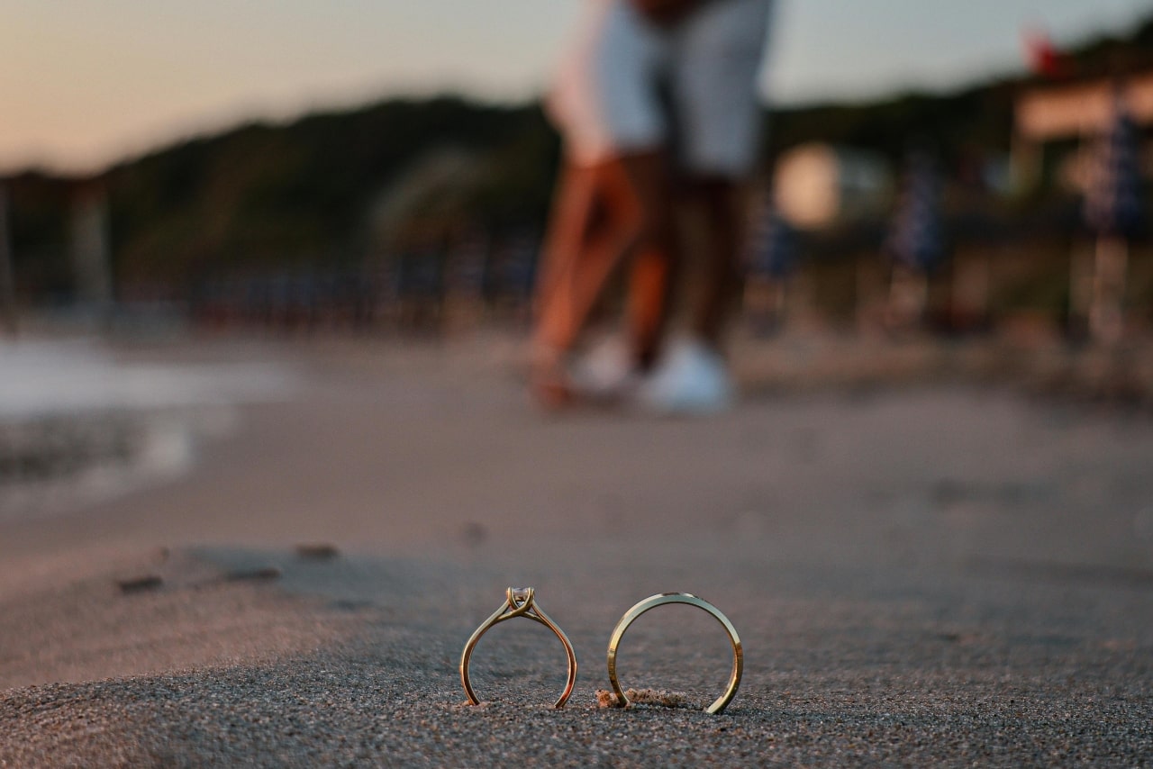 A simple engagement ring and wedding band displayed upright in beach sand, a newly engaged couple embracing in the background.