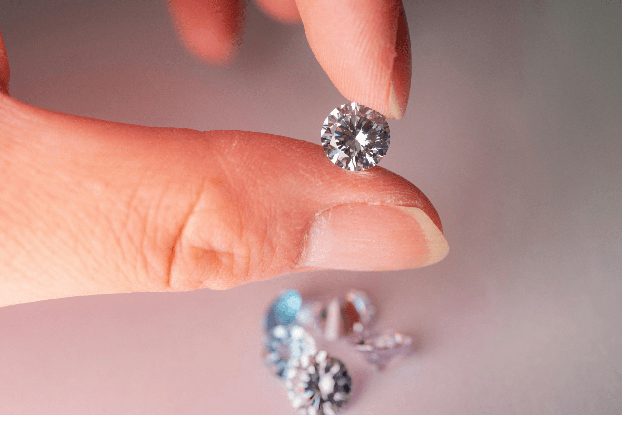 A close-up of a woman’s thumb and index finger carefully holding a stunning round cut diamond, with similar diamonds in the background.