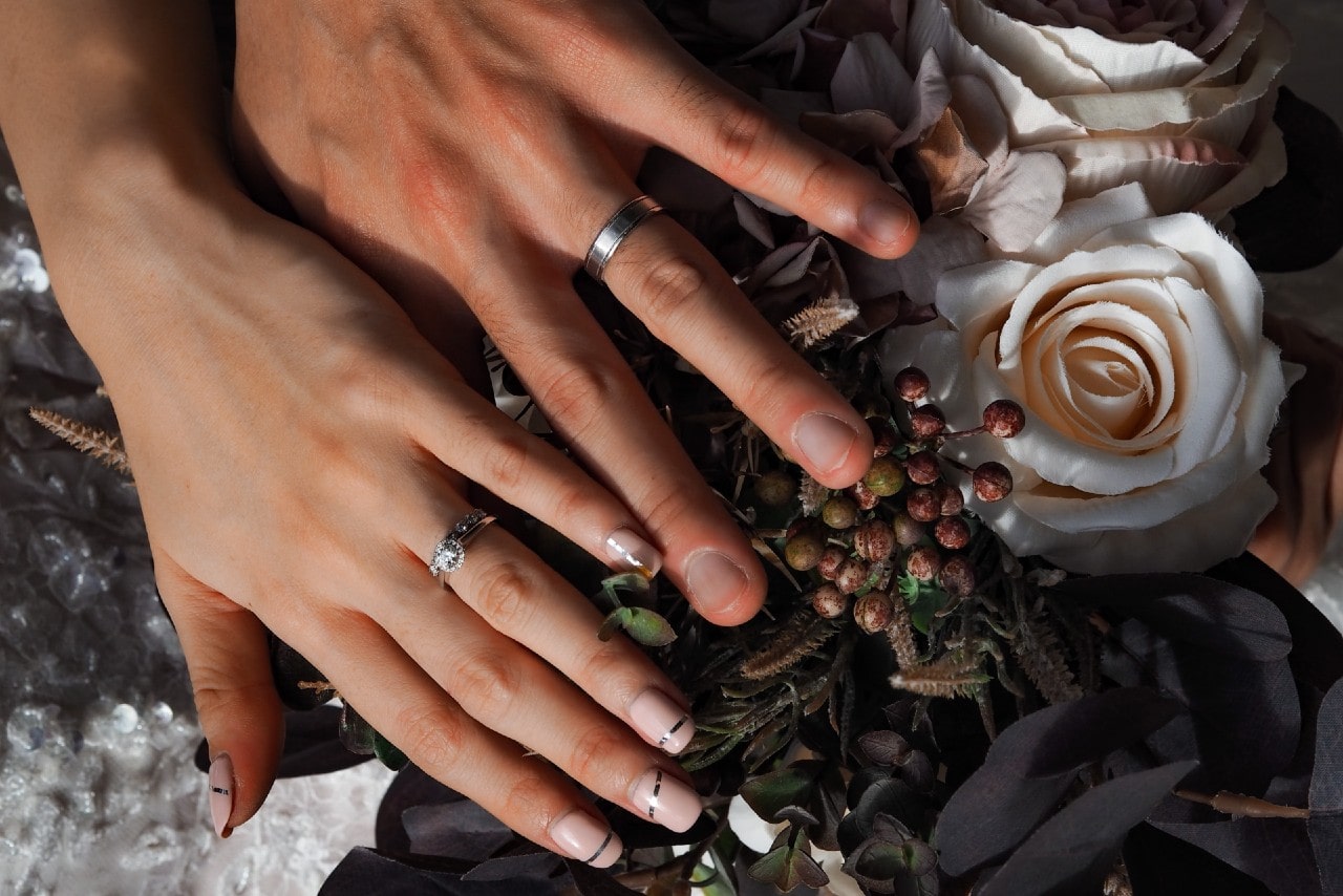 Close-up of two hands with wedding rings resting on a floral bouquet.