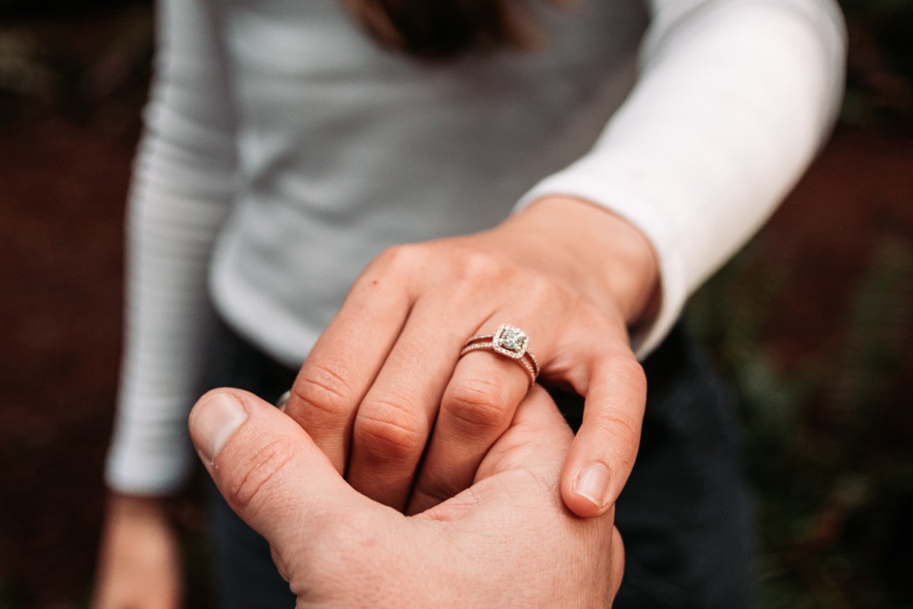 A close up of a halo double row rose gold engagement ring on the woman's finger against the blurry background of her white longsleeve and grey jeans.