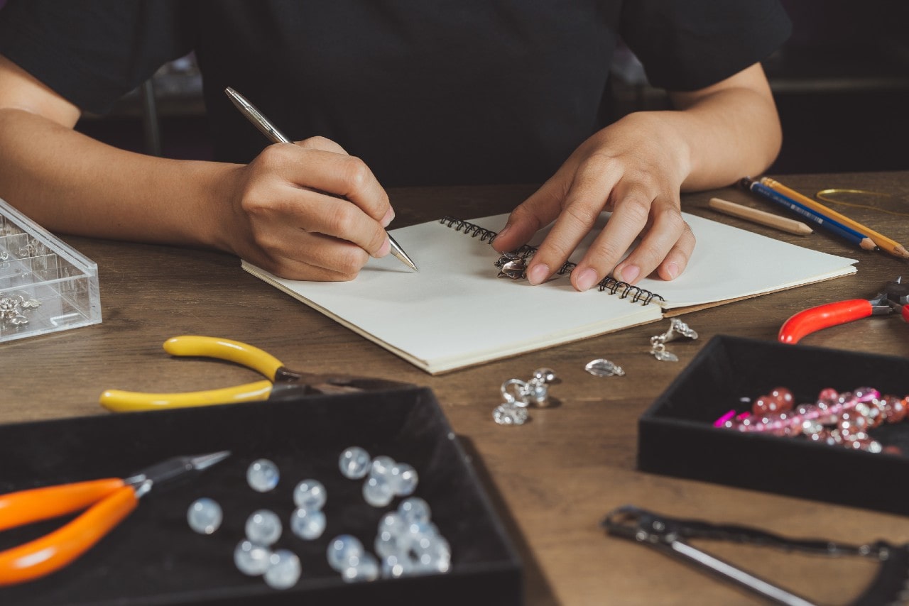 Close up of jeweler&rsquo;s hands sketching jewelry designs in a notebook on a wooden table, surrounded by beads, pliers, and tools