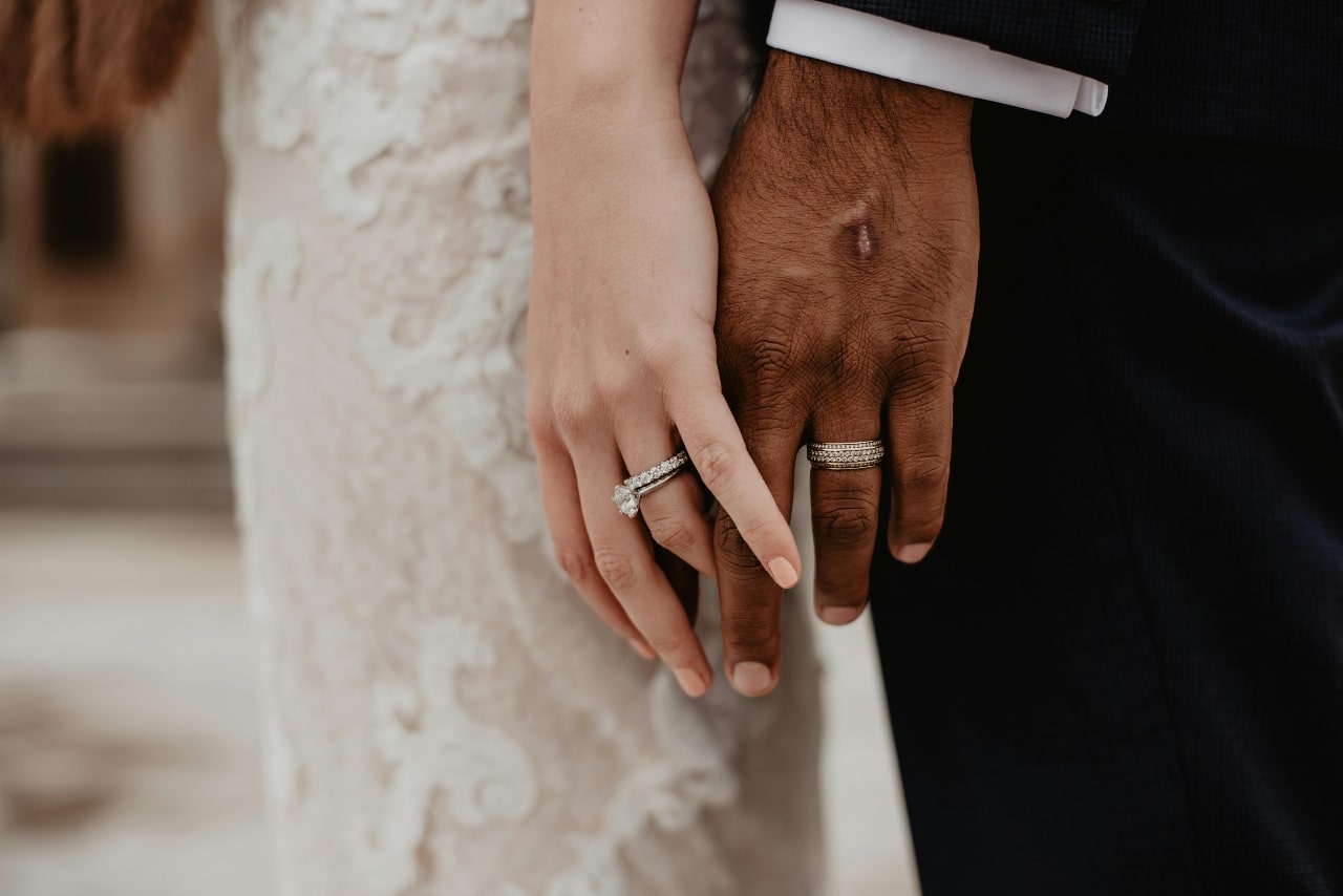 A close-up of newlyweds holding hands, their wedding rings glimmering against the bride&rsquo;s lace dress and groom&rsquo;s dark suit.