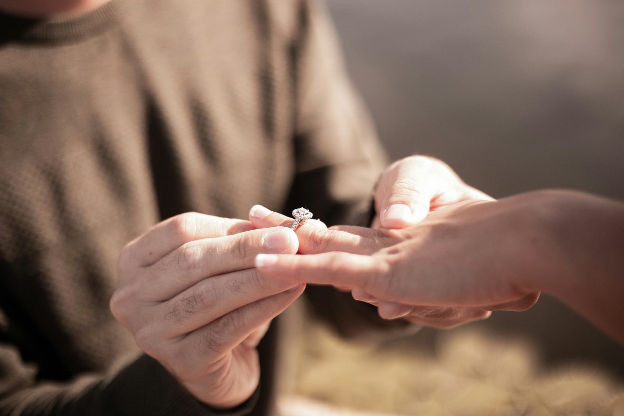 Close-up of a person in a sweater placing a ring on another’s finger, capturing a romantic proposal against a softly blurred background.