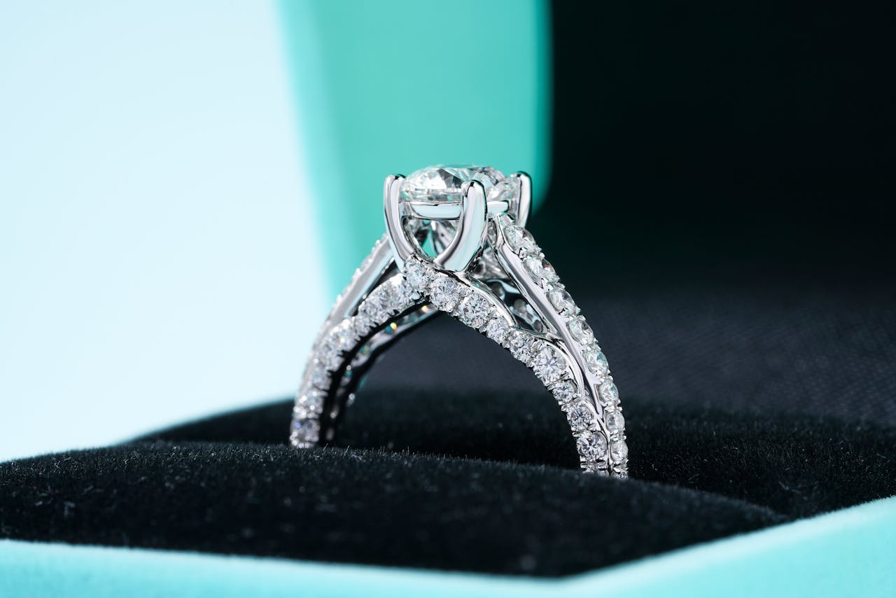 Elegant diamond ring with intricate band design in a black velvet box against a turquoise background.