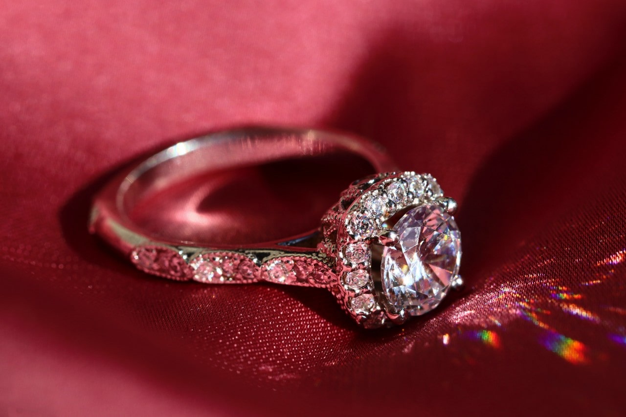 A sparkling diamond ring with a large central stone and smaller surrounding diamonds rests on luxurious red fabric.