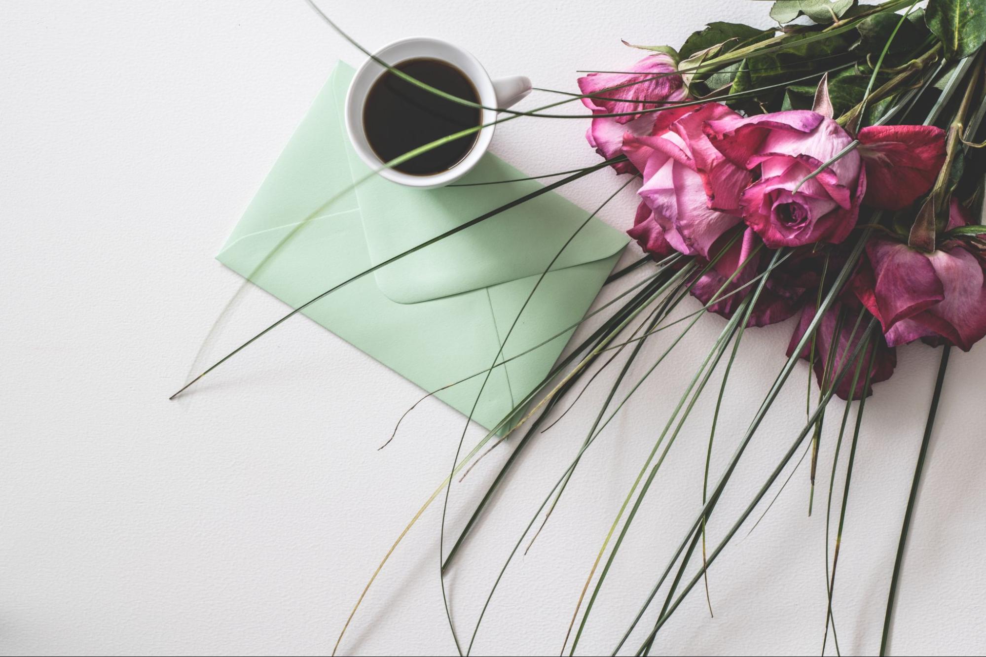 A table top with a light green envelope, a cup of black coffee, and some pink roses laid on top of it.