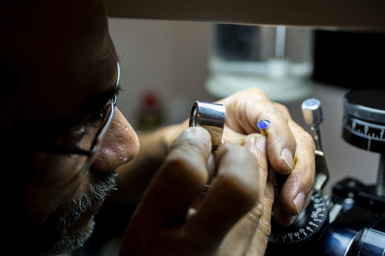 A jeweler closely examines a blue gemstone with a magnifying tool, focusing intently under soft lighting.