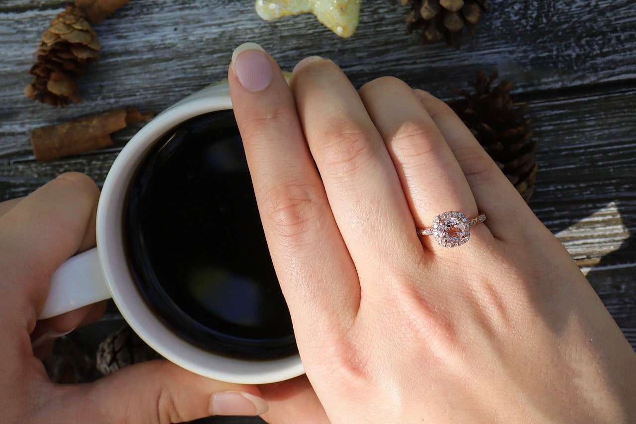 A close-up of a hand half covering a cup of coffee, showcasing white gold halo diamond engagement ring, against a wooden table surface.