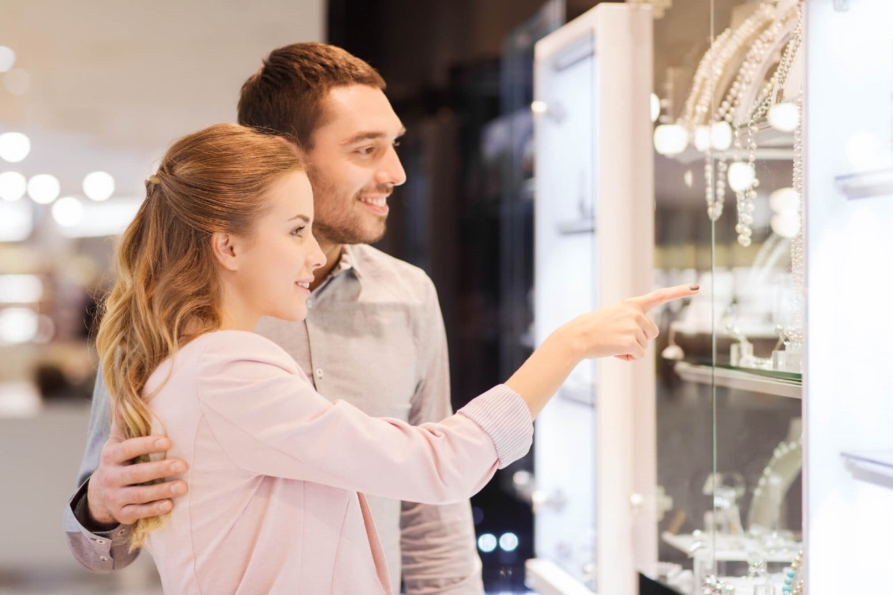 A couple standing in front of a jewelry shop window with a woman pointing at it, while a man smiling and embracing her.