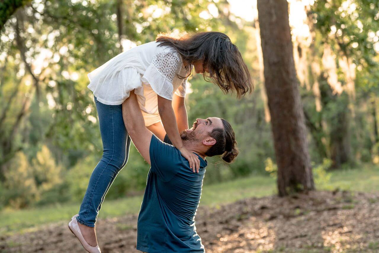 A man joyfully lifts a woman in a playful embrace in a sunlit forest, with trees and greenery in the background.