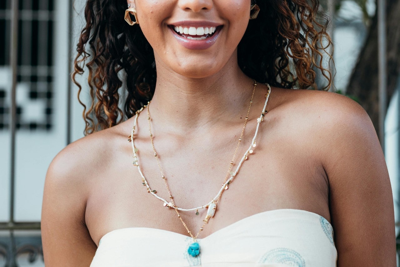 A smiling woman in a white top wearing layered summer necklaces.