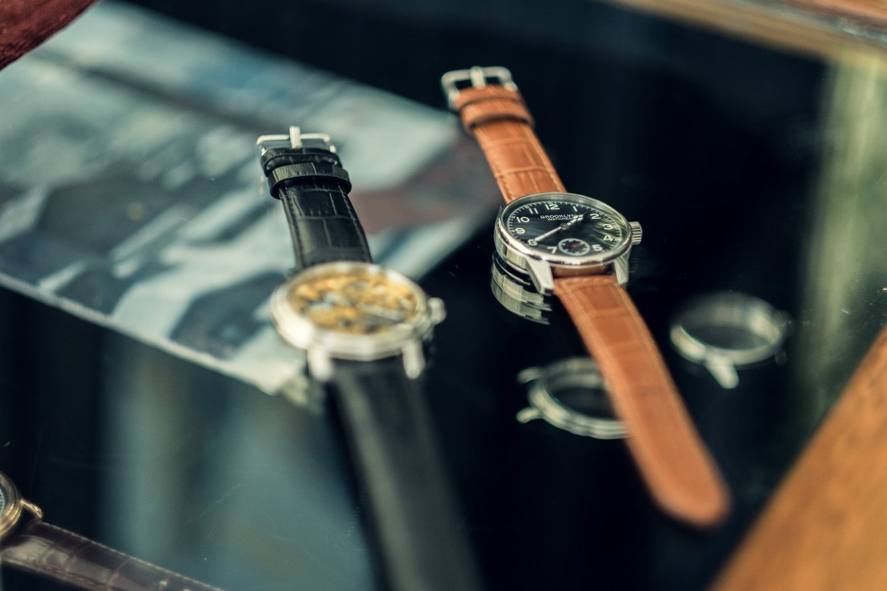 A close-up of two luxury watches with leather bands laying on top of a glass surface, with a third watch off to the side.