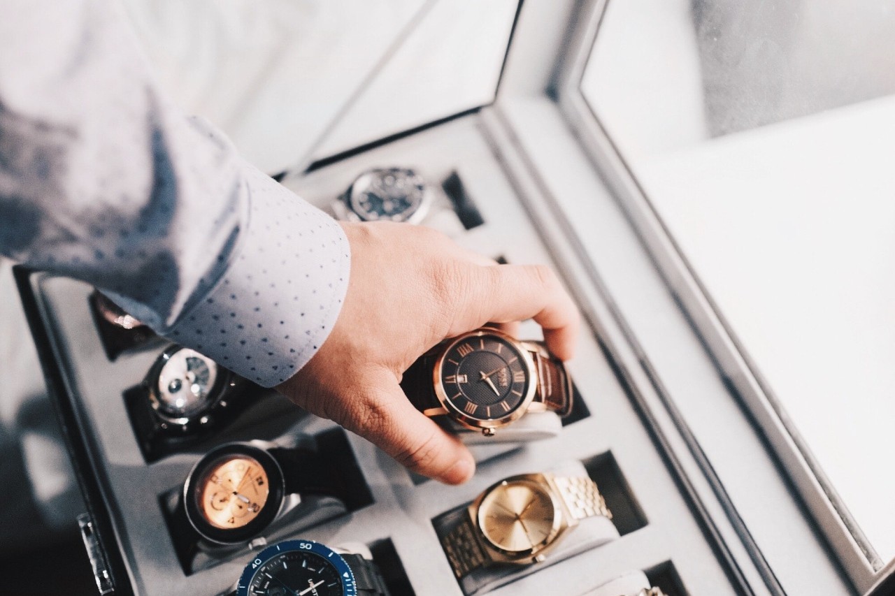 A top-down view of a man in a dress shirt selecting a bronze-toned luxury watch from a case securely housing multiple timepieces.
