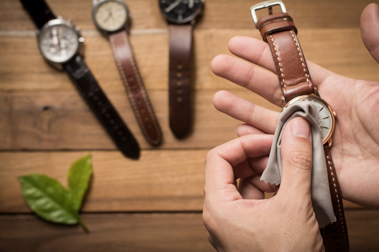 A close-up of man’s hands as he cleans the dial of a luxury watch, with three pristine luxury watches laying on a wooden surface in the background.
