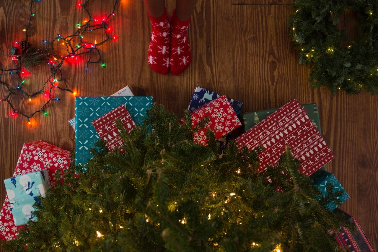 Overhead view of a festive scene with colorful wrapped gifts under a lit Christmas tree, while a person in red snowflake socks stands on a wooden floor beside glowing string lights.