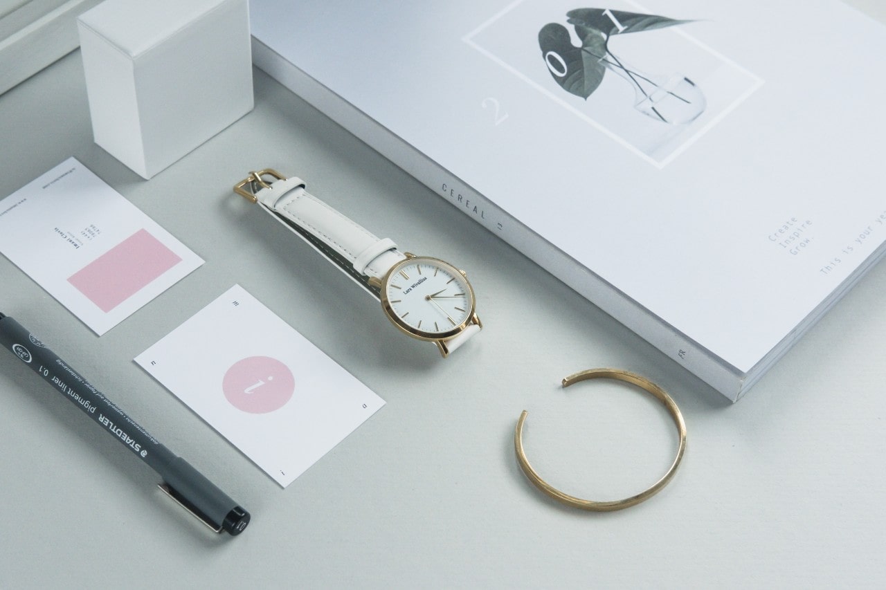 Minimalist flat lay of a white watch, gold bracelet, pen, cards with pink elements, and a stylish magazine on a light surface.