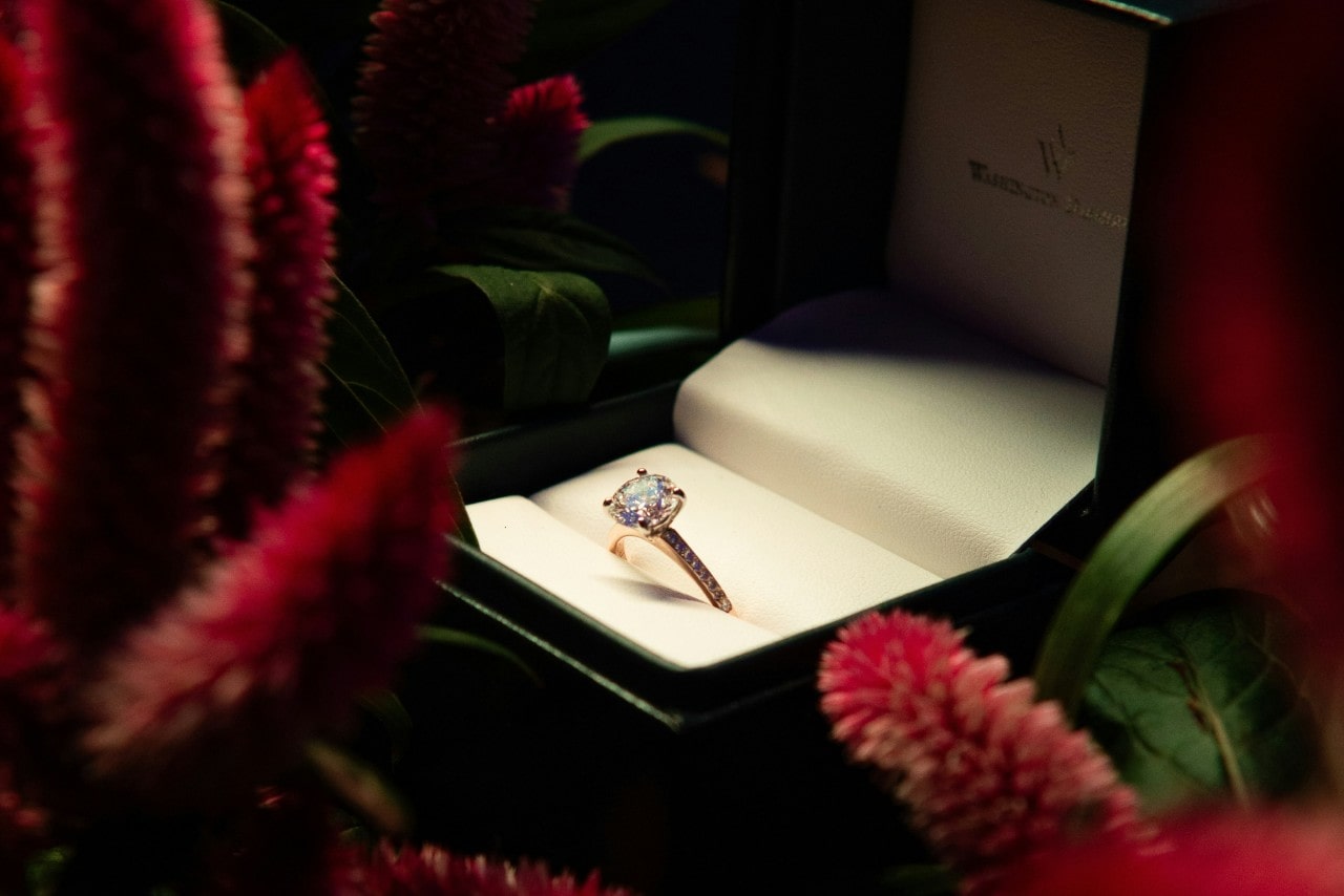 a side stone engagement ring in a green ring box surrounded by flowers