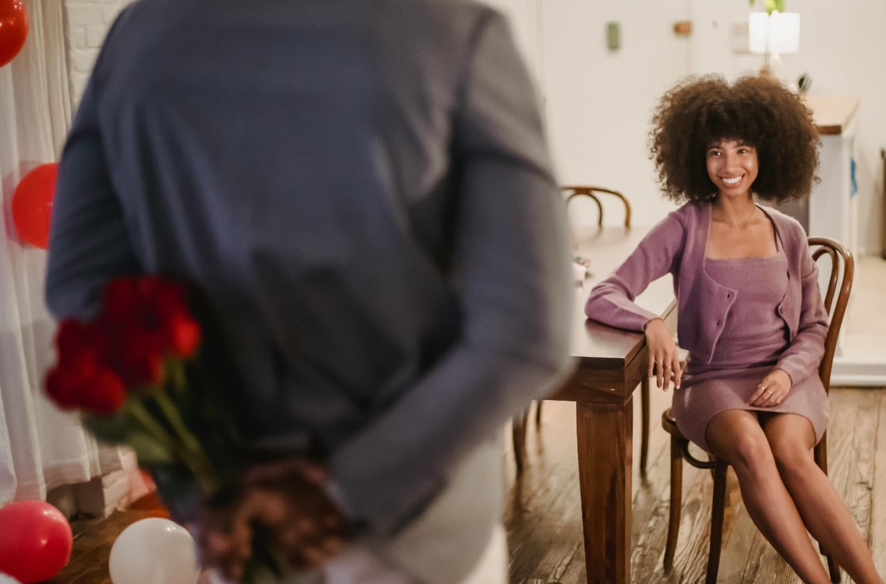 A smiling woman in a purple outfit sits at a decorated table while someone hides a bouquet of red roses behind their back