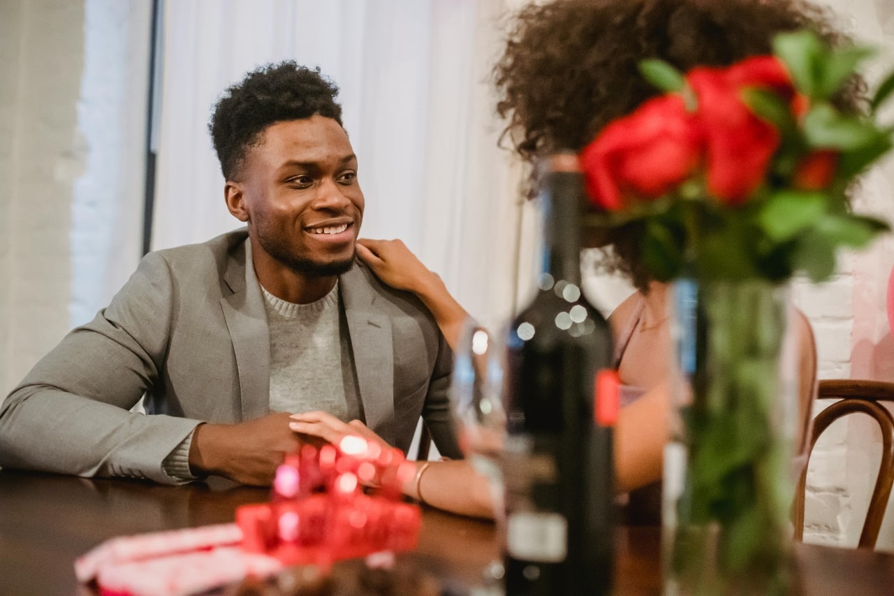 Couple sits at the table with a glass of wine and vase with red roses engaging in a romantic conversation