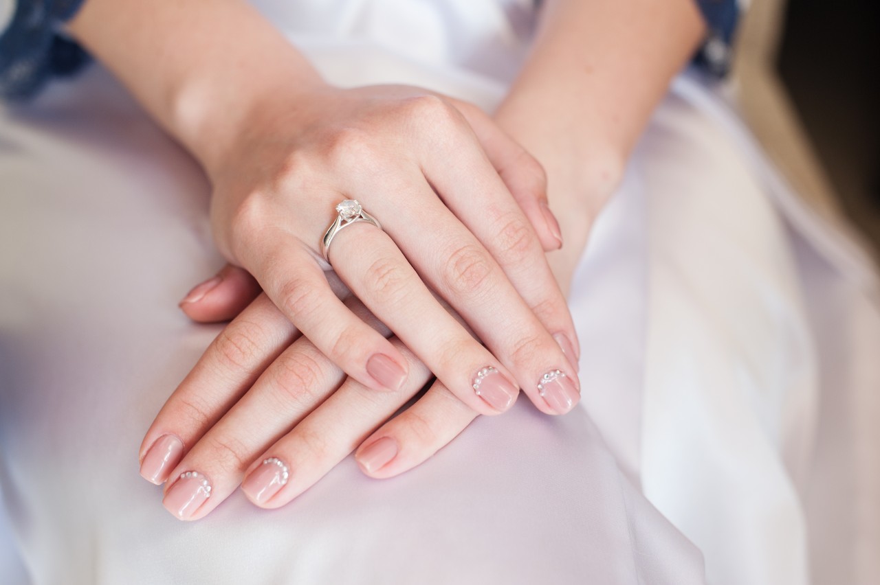 Close-up of a bride’s hands resting on a white fabric with a sparkling ring adorning one finger, and nails have a delicate manicure with pink polish and subtle designs.