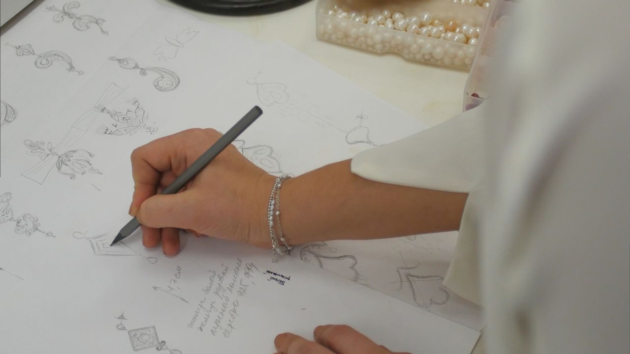 Close-up of jeweler’s hand sketching intricate jewelry designs with a pencil on white paper on a desk with scattered pearls.