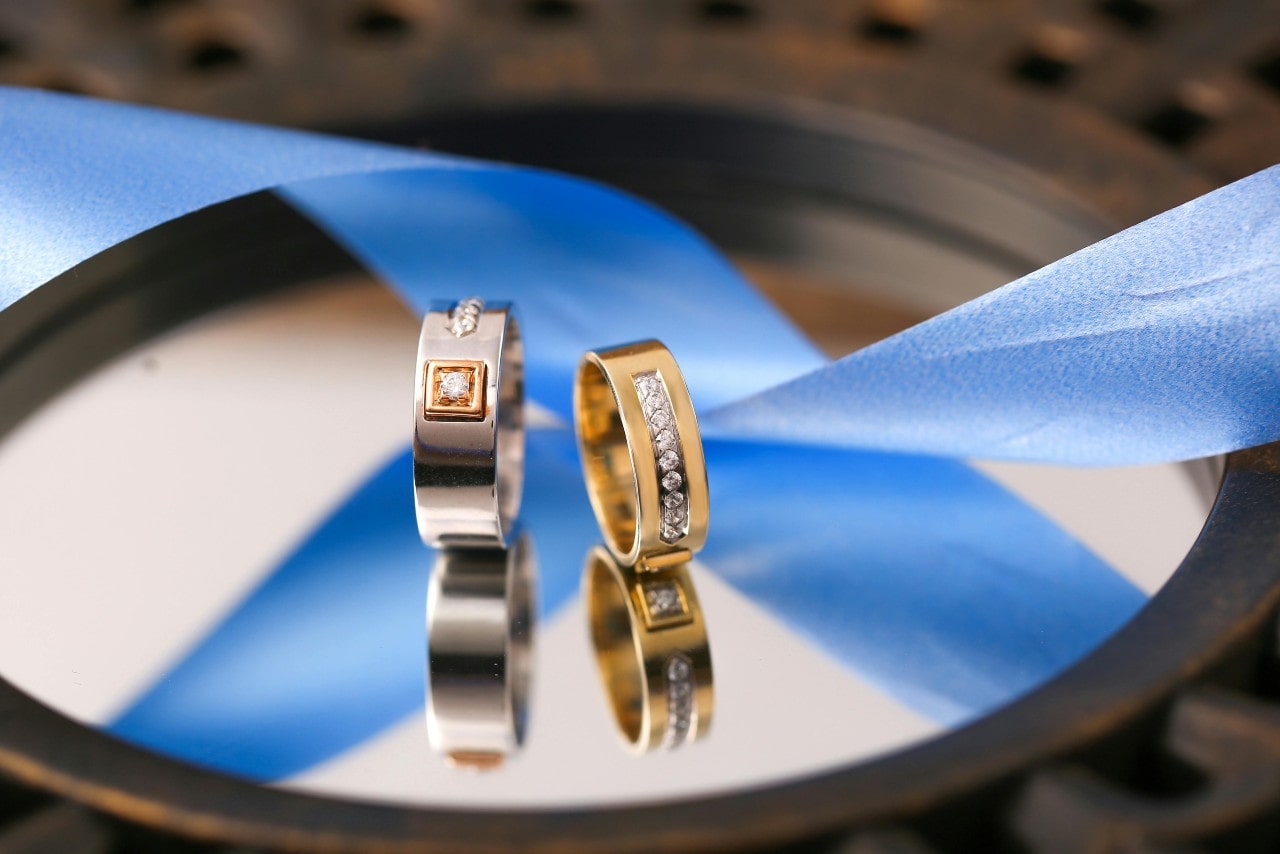 Two diamond wedding bands in white and yellow gold rest on a reflective surface beside a blue ribbon.