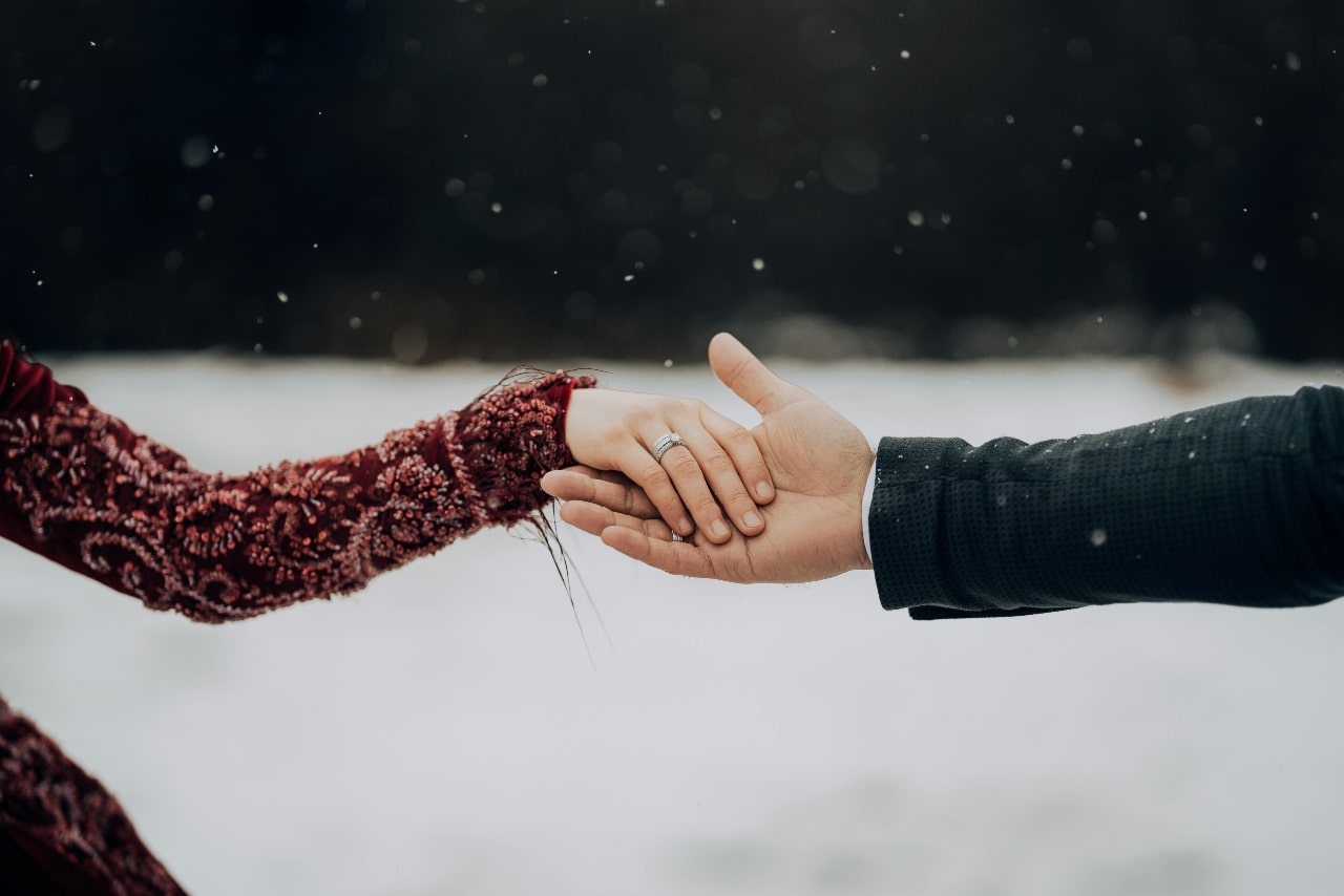 A close up of two hands in front of the snowy backdrop, showcasing white gold wedding band and an engagement ring.