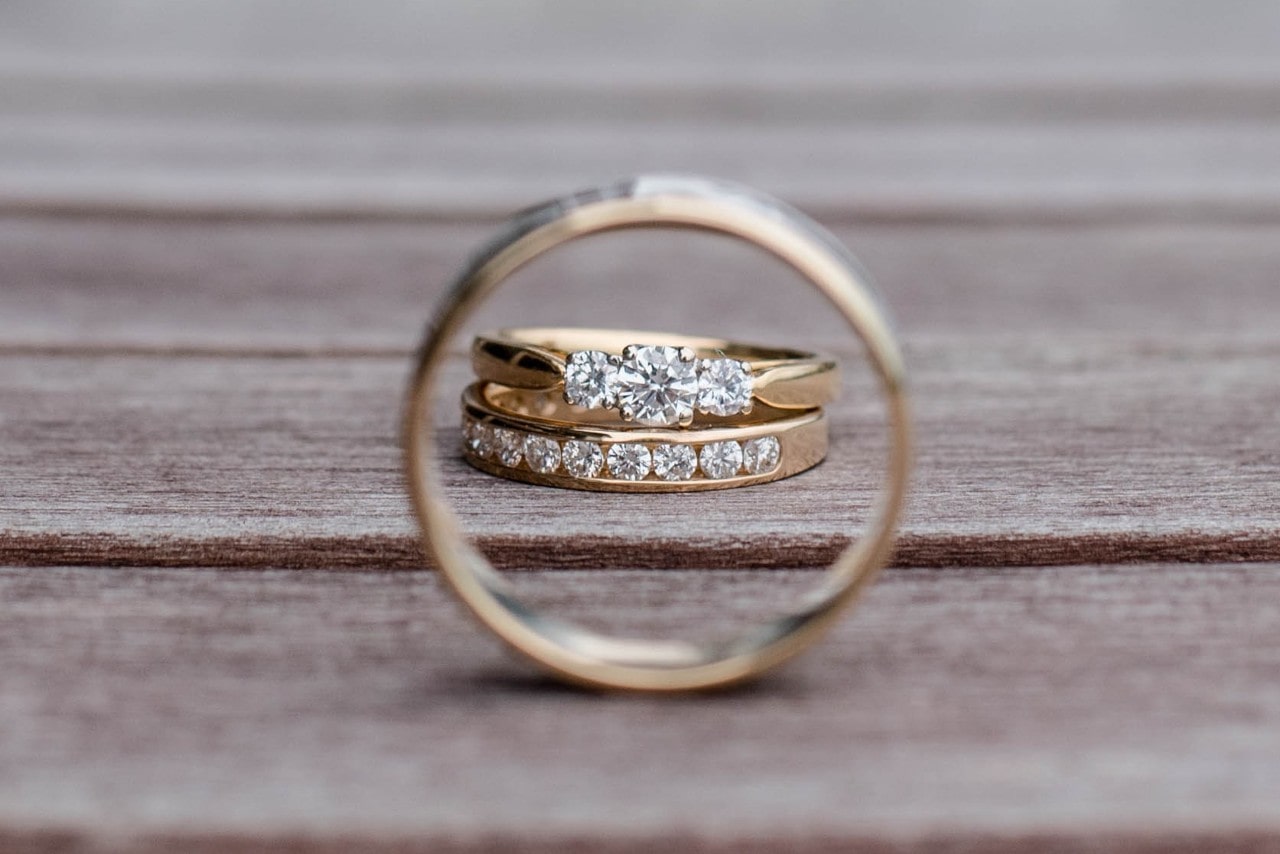 Two diamond-studded gold rings are framed through another band, resting on a wooden surface in a romantic and symmetrical composition.