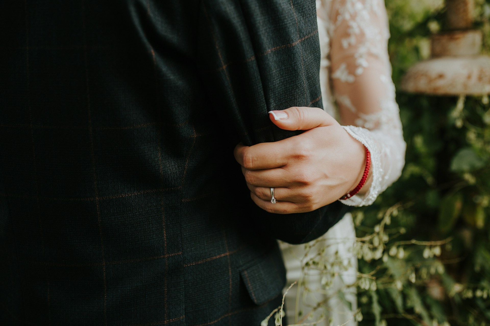 A woman’s hand in a lace sleeve rests on a man's suit-clad arm, showcasing a white gold diamond wedding band against a blurry green background.