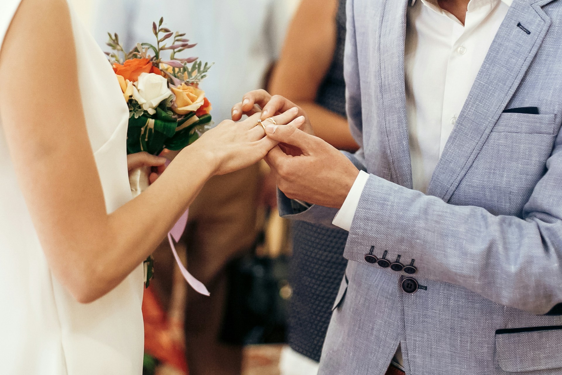 A close-up of a groom in a grey suit putting a yellow gold wedding band onto the bride's hand, while she holds a bouquet of roses in the other hand.