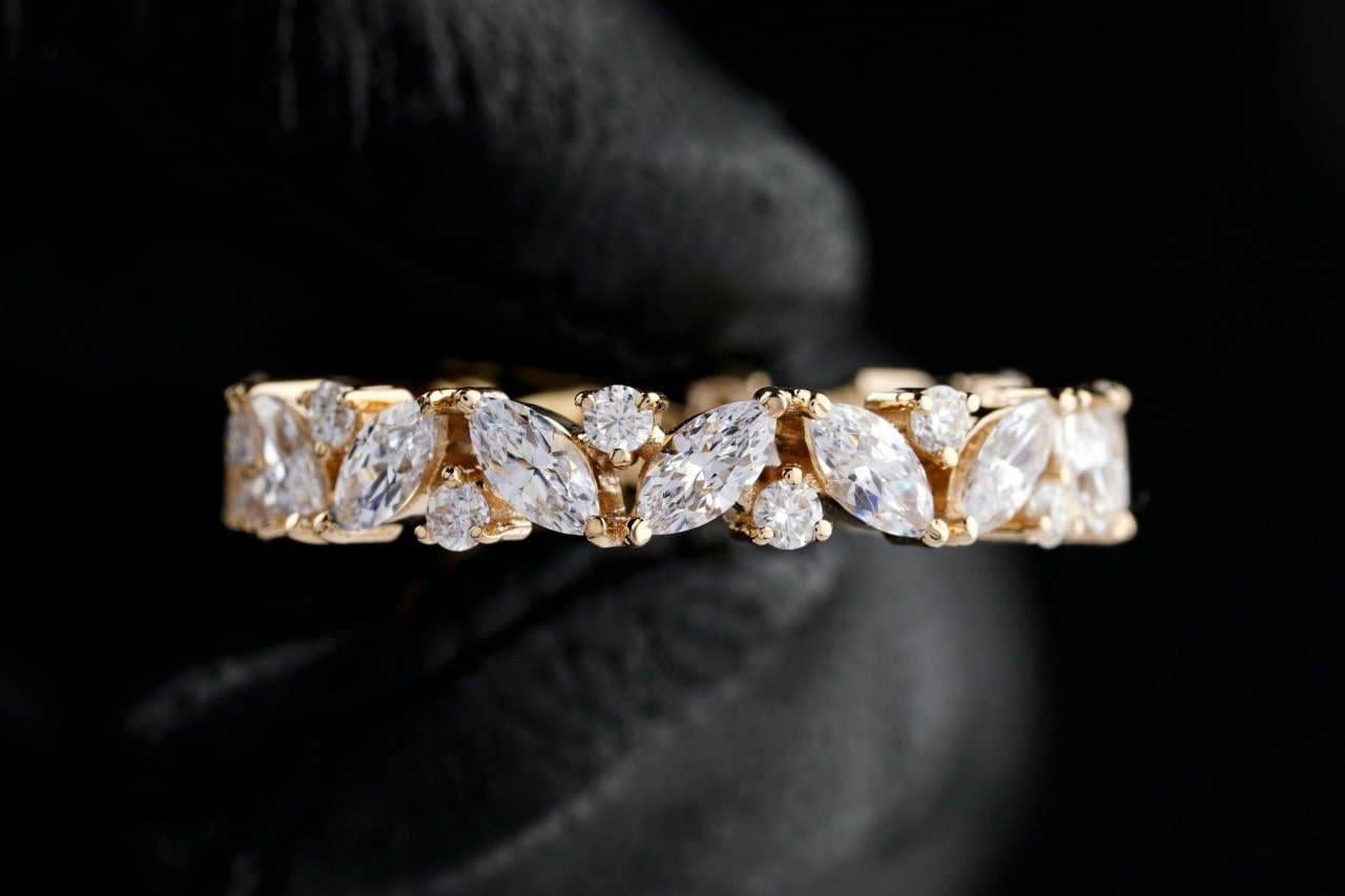 A yellow gold wedding band with marquise and round diamonds held by hands in black gloves.