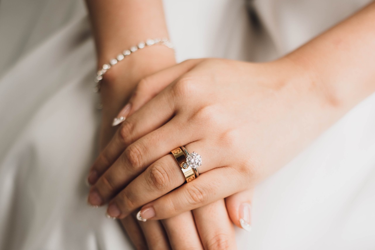 A bride’s hands with French manicure, showcasing a solitaire diamond engagement ring and yellow gold wide wedding band with a single embedded diamond on top of her wedding dress.
