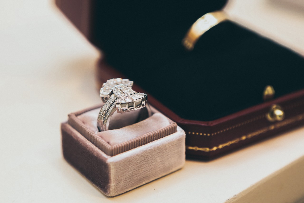 A white gold diamond ring in a velvet box on a table, with a blurred gold wedding band in a black box nearby.