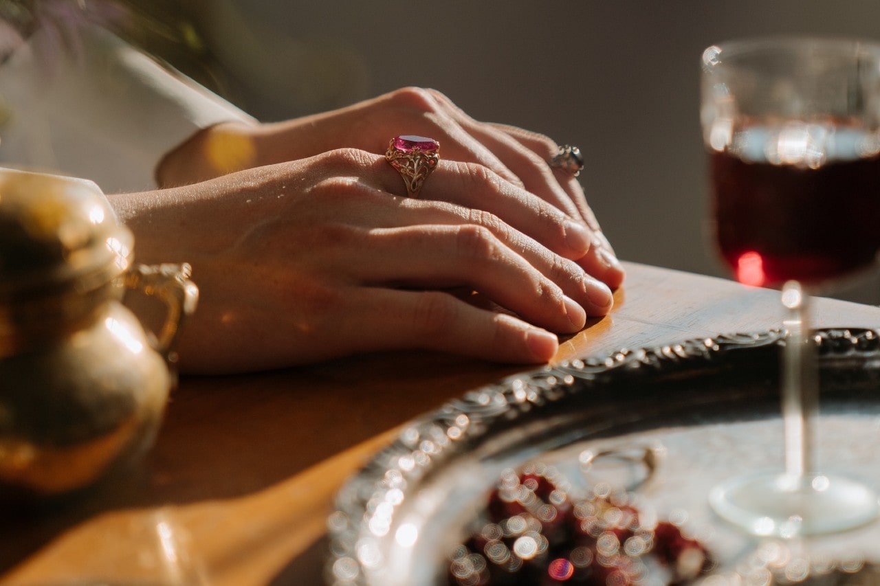 A close up of a woman’s hand on a table with glass of wine, showcasing yellow gold ring with massive pink stone.